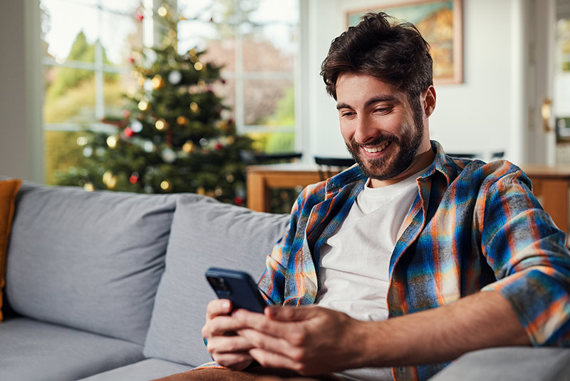 Hombre sentado en el sofá hablando por el móvil con el árbol de Navidad de fondo.